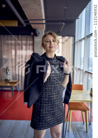 attractive woman in a black business style mini dress throws a jacket over her shoulders in a modern coworking space with panoramic windows and a black ceiling 113146932