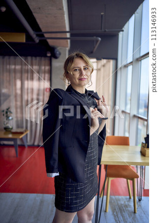 attractive woman in a black business style mini dress throws a jacket over her shoulders in a modern coworking space with panoramic windows and a black ceiling 113146933