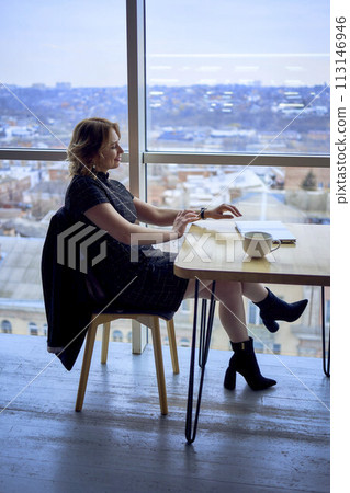 stunning mature woman at a table in a cafe drinking coffee near panoramic windows with a view of the city 113146946