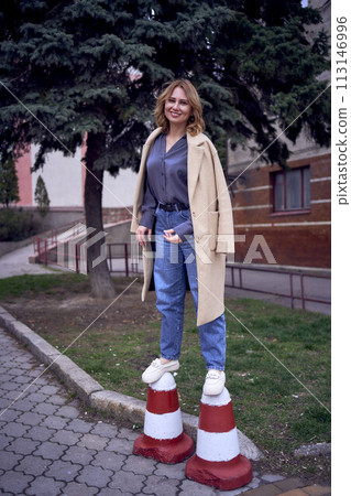 Stylish Woman Posing Casually on Traffic Cones Outside Urban Building at Dusk Stylish Woman Posing Casually on Traffic Cones Outside Urban Building at Dusk 113146996