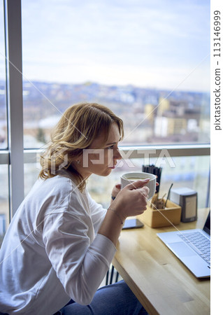 young blonde woman drinking coffee and working on a laptop in a cafe with panoramic windows and a view of the city from above 113146999