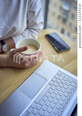 middle age woman with blond hair drinks coffee and works at a laptop in a coworking space with panoramic windows and a view of the city from above, details laptop, coffee, table 113147014