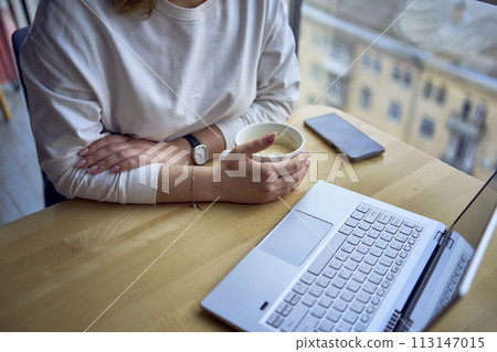 middle age woman with blond hair drinks coffee and works at a laptop in a coworking space with panoramic windows and a view of the city from above, details laptop, coffee, table 113147015