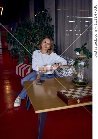 beautiful middle age woman drinking coffee in a beautiful cafe with greenery and light panels in the background beautiful middle age woman drinking coffee in a beautiful cafe with greenery and light panels in the background 113147036