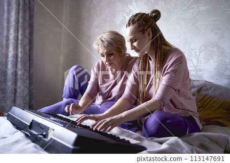 A 60-year-old mother and a 40-year-old daughter play the keyboard together on the bed at home 113147691