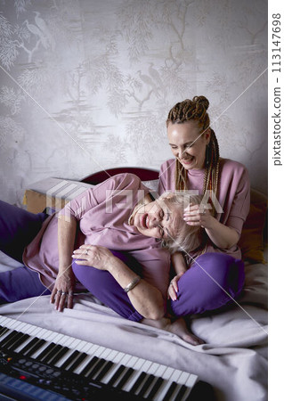 A 60-year-old mother and a 40-year-old daughter play the keyboard together on the bed at home 113147698