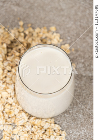 Oat milk in a glass among dry oatmeal flakes on a gray background. Oat milk in a glass among dry oatmeal flakes on a gray background. 113147699