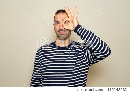 Bearded Hispanic man in his 40s wearing a striped sweater smiling while making an ok gesture over his eye in approval, isolated on beige studio background. 113148982