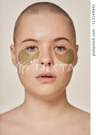 Closeup studio portrait of young Caucasian woman with shaved head and collagen eye patches looking at camera 113149943