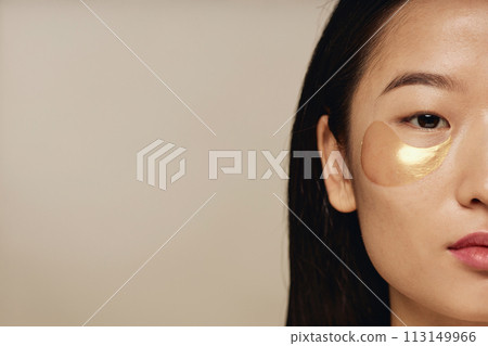 Crop half face closeup studio shot of young Asian woman with collagen patches under eyes looking at camera, copy space Crop half face closeup studio shot of young Asian woman with collagen patches under eyes looking at camera, copy space 113149966