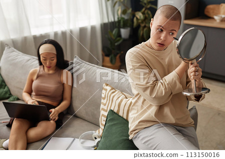 High angle view of young Caucasian woman applying mascara while her Asian girl friend working on laptop in living room at home High angle view of young Caucasian woman applying mascara while her Asian girl friend working on laptop in living room at home 113150016