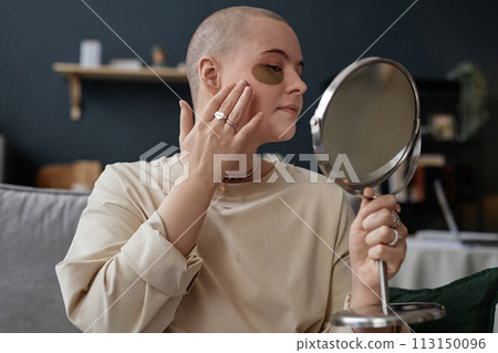 Portrait of young Caucasian woman with undereye patches on face holding mirror applying nourishing cream on skin Portrait of young Caucasian woman with undereye patches on face holding mirror applying nourishing cream on skin 113150096