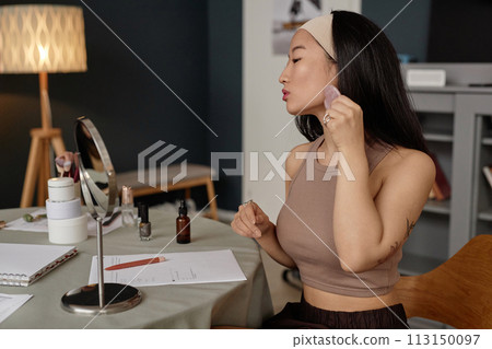 Medium shot of young Asian woman sitting at table looking into mirror doing gua sha facial massage Medium shot of young Asian woman sitting at table looking into mirror doing gua sha facial massage 113150097