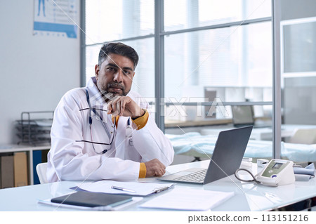 Portrait of senior Middle Eastern doctor sitting at workplace in medical clinic and looking at camera with confidence copy space 113151216
