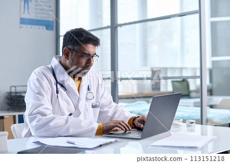 Portrait of senior Middle Eastern doctor using laptop while sitting at desk in medical clinic copy space 113151218
