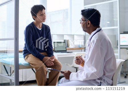 Portrait of young boy listening to doctor while sitting on cot in medical clinic copy space 113151219