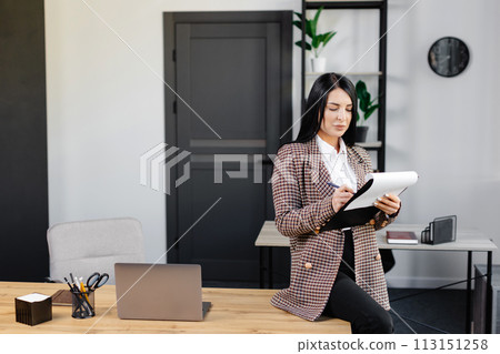 Positive woman sitting at office table writing notes on paper while working on project in modern workplace Positive woman sitting at office table writing notes on paper while working on project in modern workplace 113151258