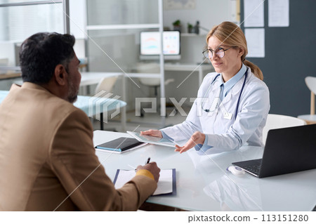 Portrait of mature female doctor consulting Indian man in medical clinic and smiling friendly 113151280