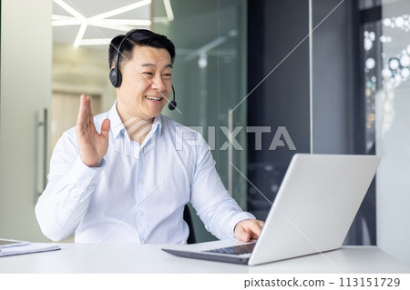 Professional Asian businessman in a white shirt, engaged in a friendly video call with a headset on, waving inside a modern office setting. 113151729