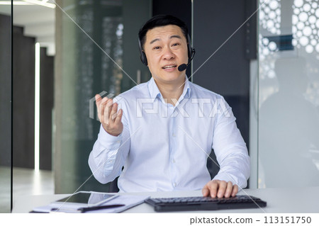 Professional Asian man in a business setting making a call with a headset, engaging with the camera. Professional Asian man in a business setting making a call with a headset, engaging with the camera. 113151750