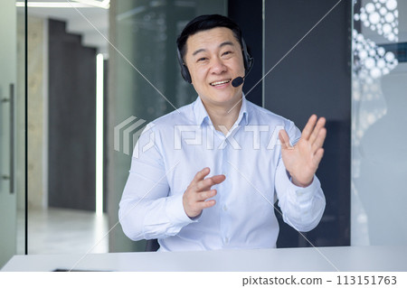 Confident Asian man in a white shirt, with a headset, is engaging in a business video call inside a modern office setting, looking at the camera. Confident Asian man in a white shirt, with a headset, is engaging in a business video call inside a modern office setting, looking at the camera. 113151763