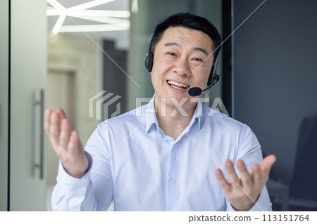 Professional Asian businessman in an office setting actively participating in a video call with a headset on. 113151764