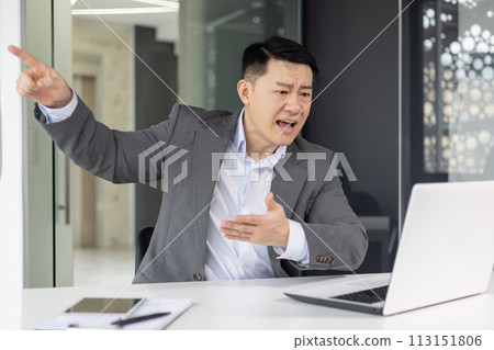 Frustrated Asian businessman gesturing while having a heated video call in a modern office setting. Frustrated Asian businessman gesturing while having a heated video call in a modern office setting. 113151806