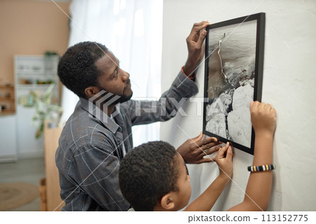 Side view portrait of African American father and son hanging picture on wall together after moving in new house Side view portrait of African American father and son hanging picture on wall together after moving in new house 113152775