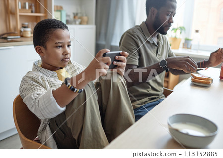 Portrait of teenage African American boy using smartphone at kitchen table during breakfast with family copy space Portrait of teenage African American boy using smartphone at kitchen table during breakfast with family copy space 113152885