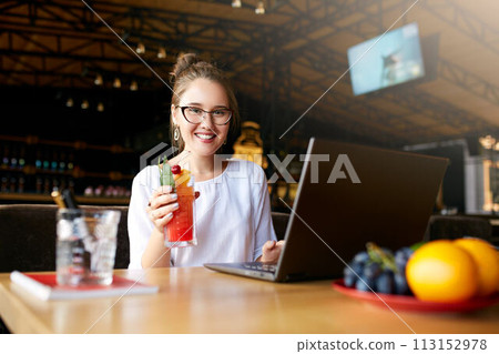 Smiling mixed race woman with cocktail in hand works with laptop. Businesswoman in glasses drinks juice for body hydration while working. Attractive designer quench thirst. Healthy lifestyle theme. Smiling mixed race woman with cocktail in hand works with laptop. Businesswoman in glasses drinks juice for body hydration while working. Attractive designer quench thirst. Healthy lifestyle theme. 113152978