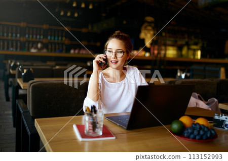 Young freelancer working with laptop talking on cellphone with client in cafe. Pretty asian caucasian mixed race business woman in glasses conducts negotiations via phone call. Multitasking concept. Young freelancer working with laptop talking on cellphone with client in cafe. Pretty asian caucasian mixed race business woman in glasses conducts negotiations via phone call. Multitasking concept. 113152993