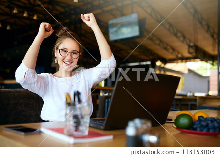 Portrait of happy young business woman celebrating success with arms up in front of laptop. Mixed race female won a lot of money in lottery prize, raised arms with fists. Freelancer finished project. 113153003