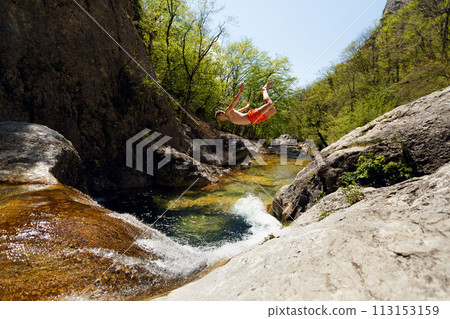 Young Man Jumping From Cliff Into Water of Mountain River 113153159