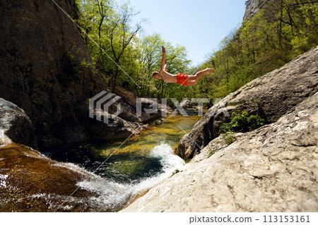 Young Man Jumping From Cliff Into Water of Mountain River 113153161