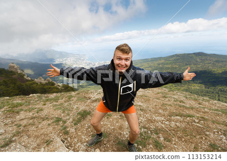 Happy smiling man stands on a top of the mountain with open hands and strong expession looking at camera with summer mountain landscape on hiking trip 113153214