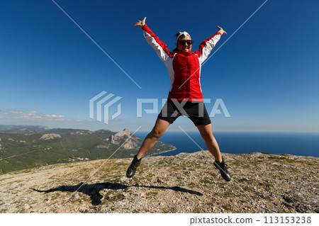 Happy woman jumps on a peak of a mountain in summer time in mountains enjoying the climb with beautiful rocky and sea background 113153238