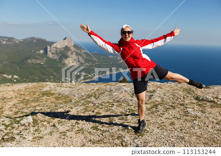 Happy woman jumps on a peak of a mountain in summer time in mountains enjoying the climb with beautiful rocky and sea background 113153244