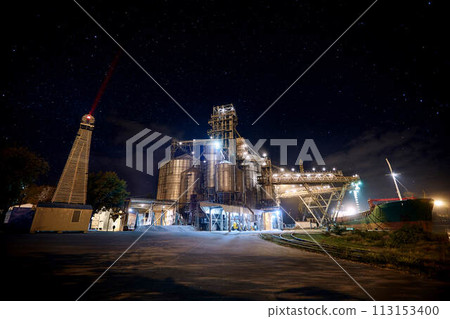 Grain terminal at seaport with starry sky on background. Cereals bulk transshipment from road transport to vessel at night. Loading grain crops on ship from elevators at the berth. Long exposure. 113153400
