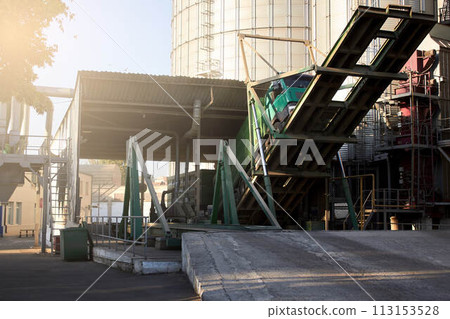 Unloading grain truck at elevator on elevating hydraulic platform unloader. Grain crops transshipment at big sea terminal at seaport. Unloading grain truck at elevator on elevating hydraulic platform unloader. Grain crops transshipment at big sea terminal at seaport. 113153528
