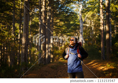 Hiker man walks in a pine yellow autumn forest. Backpacker enjoys fall landscape. Tourist wears sport sunglasses. Hiker man walks in a pine yellow autumn forest. Backpacker enjoys fall landscape. Tourist wears sport sunglasses. 113153547