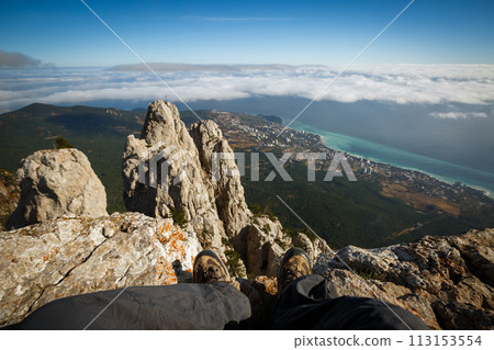 Traveler sitting on a cliff of rocky mountain summit above clouds. Point of view photo of legs in hiking boots with sea, city and mountain landscape. Ai Petri, Yalta, Crimea. 113153554