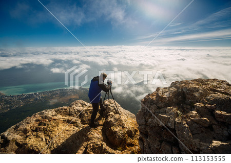 Man standing with a tripod and camera on a high mountain peak above clouds, city and sea. Professional photographer adjusting dslr settings on rocky summit. 113153555