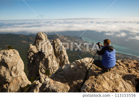 Man sitting with a tripod and photo camera on a high mountain peak above clouds, city and sea. Pro photographer adjusting dslr settings on rocky summit. 113153556