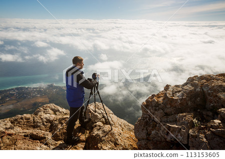 Man standing with a tripod and camera on a high mountain peak above clouds, city and sea. Professional photographer adjusting dslr settings on rocky summit. 113153605