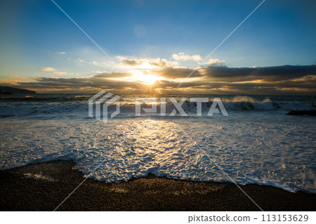 Morning photo of a sunrise above the stormy sea on empty pebble sand beach and foam. Dramatic sky with sunbeams and flares through clouds. The wave is breaking about the shore. Morning photo of a sunrise above the stormy sea on empty pebble sand beach and foam. Dramatic sky with sunbeams and flares through clouds. The wave is breaking about the shore. 113153629