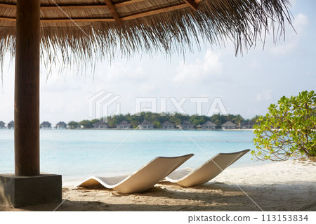 Two modern beach chairs on the beach under straw umbrella. Indian ocean coastline on Maldives island. White sandy shore and calm sea. Travel and vacation concept. Two modern beach chairs on the beach under straw umbrella. Indian ocean coastline on Maldives island. White sandy shore and calm sea. Travel and vacation concept. 113153834
