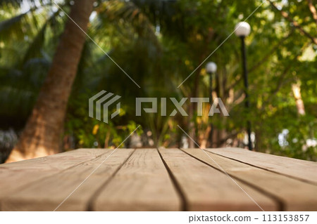 Empty top of natural wooden table for product placement and display in open shade. Coconut palm trees and green tropical plants on background. Maldives 113153857