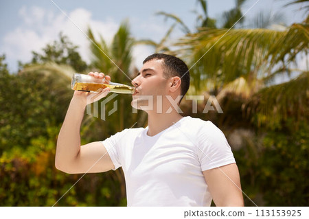 Young man drinking beer on the tropical beach with coconut palm trees on background. Caucasian male with a bottle of lemonade beverage quenches thirst. Young man drinking beer on the tropical beach with coconut palm trees on background. Caucasian male with a bottle of lemonade beverage quenches thirst. 113153925