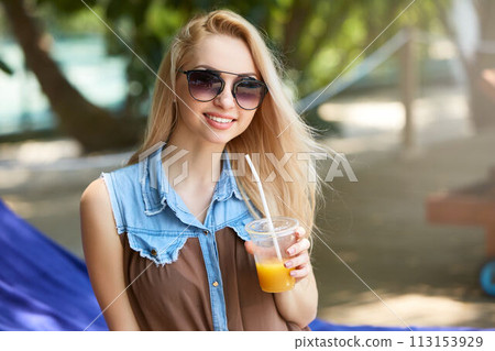 Portrait of young woman with cocktail glass chilling in the tropical sun near swimming pool on a deck chair with palm trees behind. Vacation concept 113153929