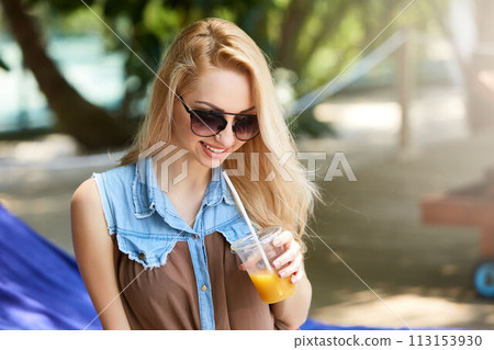 Portrait of young woman with cocktail glass chilling in the tropical sun near swimming pool on a deck chair with palm trees behind. Vacation concept Portrait of young woman with cocktail glass chilling in the tropical sun near swimming pool on a deck chair with palm trees behind. Vacation concept 113153930